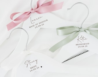 Può includere: Tre grucce in legno bianco con ganci in metallo argentato. Ogni gruccia presenta un fiocco in nastro di raso e testo stampato in nero. Il testo riporta Lauren Maid of Honour 24.08.26, Katie Bridesmaid 24.08.26 e Stacey Bride 24.08.26 con un cuore.