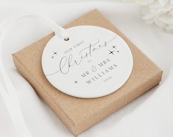 May include: A round white ceramic ornament with a white ribbon, featuring black printed text that reads Our First Christmas as Mr & Mrs Williams 2026, accented with small star symbols, resting on a square brown cardboard gift box.