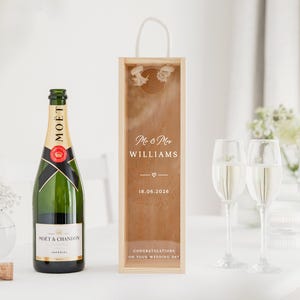 May include: A green champagne bottle with a gold label and black ribbon, beside a wooden gift box with the text "Mr & Mrs Williams" and the date "18.06.2026". Two champagne flutes and a vase of white flowers are also visible.