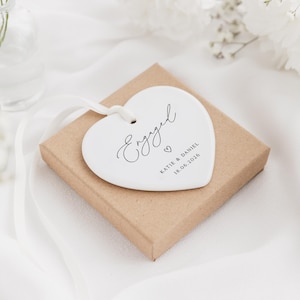May include: A white heart-shaped tag with the word "Engaged" in elegant script, along with the names "Katie & Daniel" and the date "18.06.2026". The tag is on a small brown box, with a white ribbon. The background is white fabric and flowers.