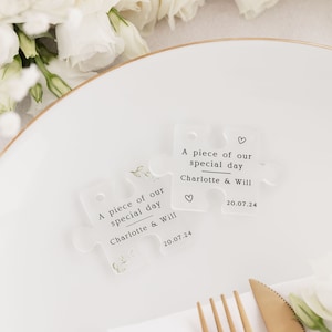 May include: Two clear acrylic puzzle pieces with the text "A piece of our special day Charlotte & Will 20.07.24" and a small heart symbol. The pieces are on a white plate with a gold rim.