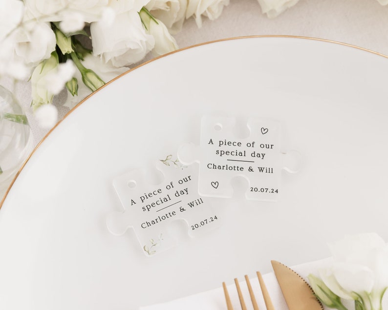 May include: Two clear acrylic puzzle pieces with the text "A piece of our special day Charlotte & Will 20.07.24" and a small heart symbol. The pieces are on a white plate with a gold rim.