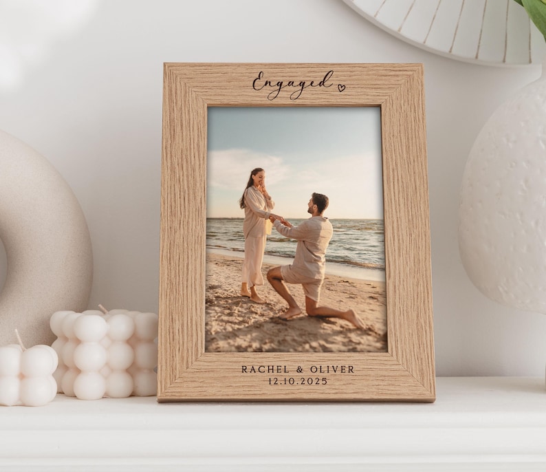 Pode incluir: Uma moldura de fotografia de madeira com uma foto de um casal na praia. A moldura &eacute; gravada com a palavra "Engaged", os nomes do casal e a data "12.10.2025".