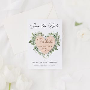 May include: A white Save the Date card with a wooden heart design. The heart is surrounded by green foliage and features the names Charlotte & Daniel and the date 12.08.27. The card also includes the text "The Willow Barn, Cotswolds" and "Formal Invitation to Follow."