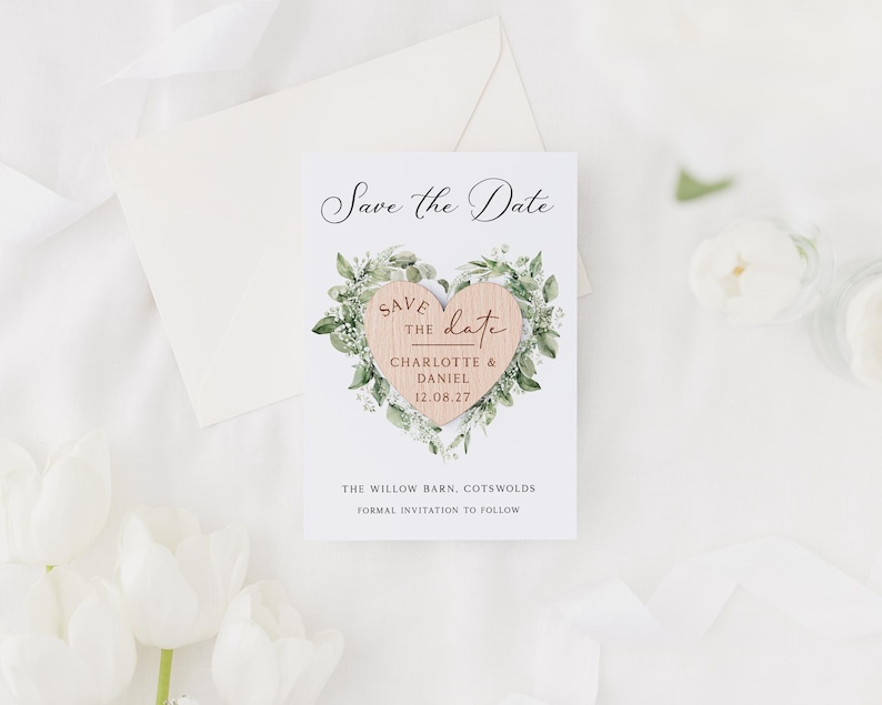 May include: A white Save the Date card with a wooden heart design. The heart is surrounded by green foliage and features the names Charlotte & Daniel and the date 12.08.27. The card also includes the text "The Willow Barn, Cotswolds" and "Formal Invitation to Follow."