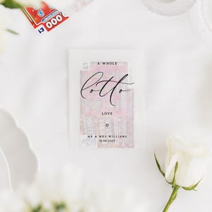 Puede incluir: Un paquete transparente con un billete de loter&iacute;a en su interior, con las palabras "A Whole Lotto Love" en escritura elegante. El paquete incluye los nombres "Mr & Mrs Williams" y la fecha "18.06.2027". Tambi&eacute;n se ve una rosa blanca y un billete de loter&iacute;a.
