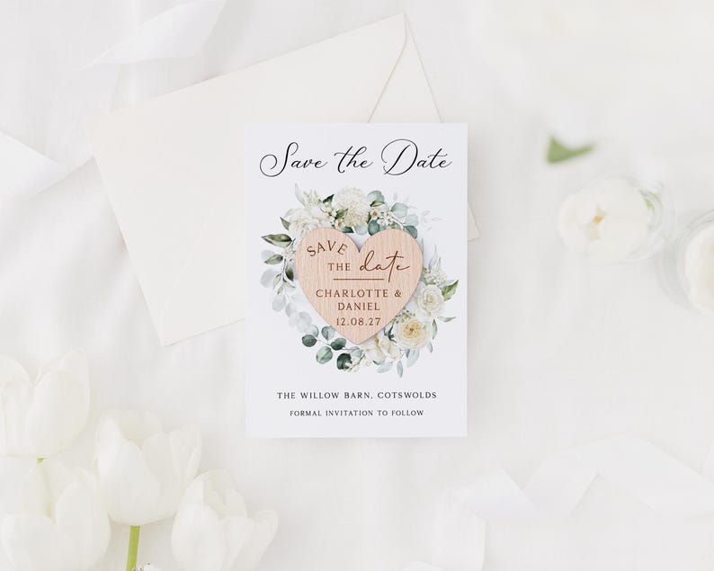 May include: A wedding announcement card with the text "Save the Date" in elegant script. The card features a wooden heart with the names "Charlotte & Daniel" and the date "12.08.27", surrounded by a floral wreath. An envelope and flowers are in the background.