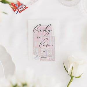 Puede incluir: Un recuerdo de boda con un billete de loter&iacute;a dentro de un sobre transl&uacute;cido. El sobre est&aacute; impreso con las palabras "Lucky in Love" en escritura elegante, junto con los nombres "Mr & Mrs Williams" y la fecha "18.06.2022". Una rosa blanca en primer plano.