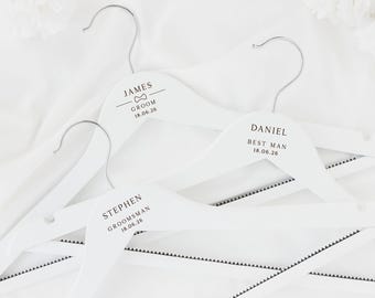 Può includere: Tre grucce in legno bianco con ganci argentati, personalizzate con nomi e ruoli in testo nero. Una recita "JAMES SPOSO 18.06.26" con un papillon. Altre dicono "DANIEL TESTIMONE 18.06.26" e "STEPHEN TESTIMONE 18.06.26".