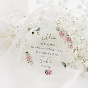 May include: A clear, circular acrylic tag with the word "Mum" in script, adorned with watercolour floral designs. The tag features a message and the date 24.08.2026, suspended by a white ribbon, and set against a backdrop of white flowers.