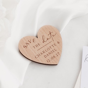May include: A wooden heart-shaped wedding announcement with the words "Save the Date" and the names "Charlotte & Daniel" along with the date "12.08.27" engraved on it. The announcement is set on a white satin background.