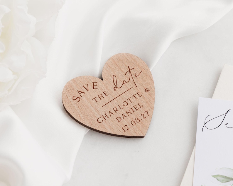 May include: A wooden heart-shaped wedding announcement with the words "Save the Date" and the names "Charlotte & Daniel" along with the date "12.08.27" engraved on it. The announcement is set on a white satin background.