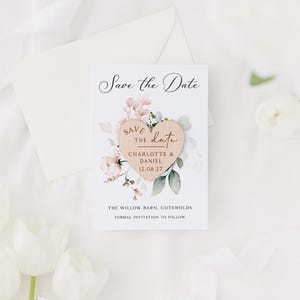 May include: A wedding announcement card with a wooden heart and floral accents. The card reads "Save the Date" and includes the names "Charlotte & Daniel" and the date "12.08.27". An envelope and flowers are in the background.