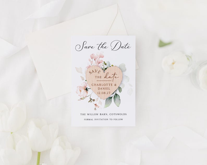 May include: A wedding announcement card with a wooden heart and floral accents. The card reads "Save the Date" and includes the names "Charlotte & Daniel" and the date "12.08.27". An envelope and flowers are in the background.