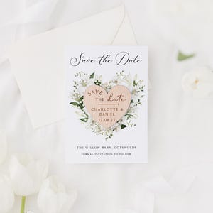 May include: A wedding announcement card with a wooden heart and floral design. The card reads "Save the Date" with the names "Charlotte & Daniel" and the date "12.08.27". Includes an envelope and white flowers.
