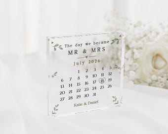 May include: Clear acrylic block with the text "The day we became MR & MRS July 2026" and a calendar highlighting July 18th. The names "Katie & Daniel" are printed at the bottom, with floral accents.