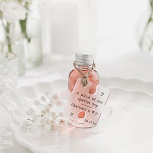 May include: A small clear glass bottle filled with a pink liquid and a white puzzle piece tag that says "A piece of our special day Charlotte & Will 20.07.24" with a heart symbol on each side of the tag.
