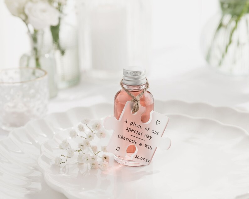 May include: A small clear glass bottle filled with a pink liquid and a white puzzle piece tag that says "A piece of our special day Charlotte & Will 20.07.24" with a heart symbol on each side of the tag.