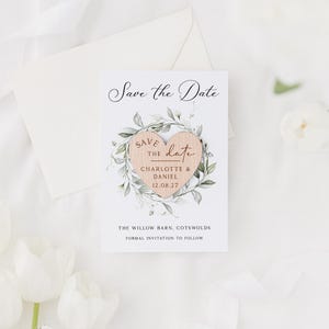 May include: A white "Save the Date" card with a wooden heart design. The heart is surrounded by green leaves and contains the names "Charlotte & Daniel" and the date "12.08.27". The card is accompanied by a white envelope and white flowers.