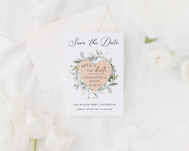 May include: A white "Save the Date" card with a wooden heart design. The heart is surrounded by green leaves and contains the names "Charlotte & Daniel" and the date "12.08.27". The card is accompanied by a white envelope and white flowers.
