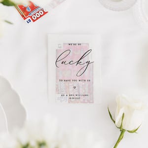Puede incluir: Un recuerdo de boda con un billete de loter&iacute;a dentro de un sobre transparente. El sobre tiene las palabras "We're so lucky to have you with us" impresas, junto con un s&iacute;mbolo de coraz&oacute;n y los nombres "Mr & Mrs Williams" y la fecha "18.06.2022".