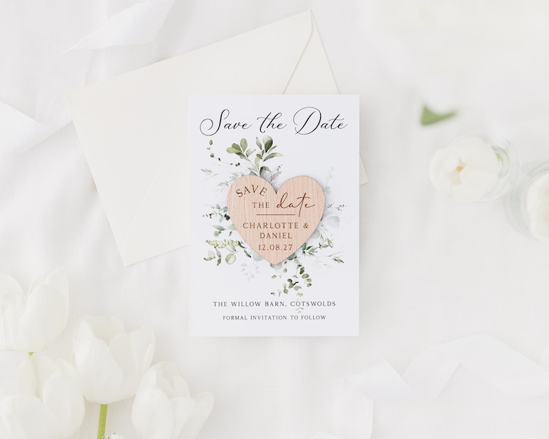 May include: A Save the Date card with a wooden heart and floral accents. The card features the names Charlotte & Daniel and the date 12.08.27. The text "Save the Date" is written in elegant script. An envelope and flowers are in the background.