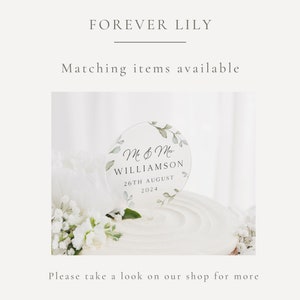 May include: A clear acrylic cake topper with a circle design featuring green leaves and the text "M & M WILLIAMSON 26TH AUGUST 2024".