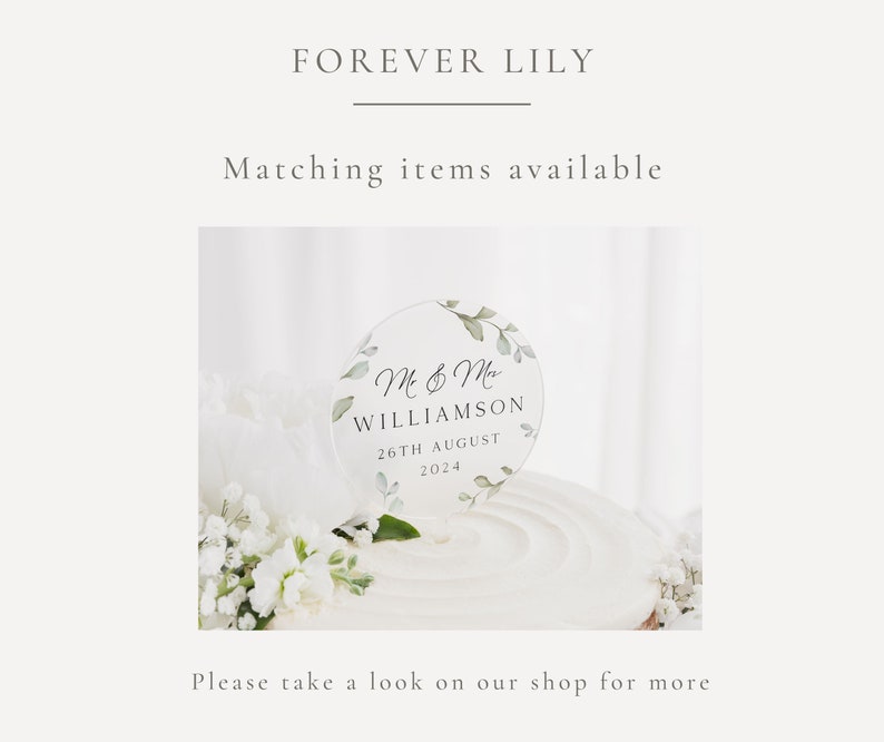 May include: A clear acrylic cake topper with a circle design featuring green leaves and the text "M & M WILLIAMSON 26TH AUGUST 2024".