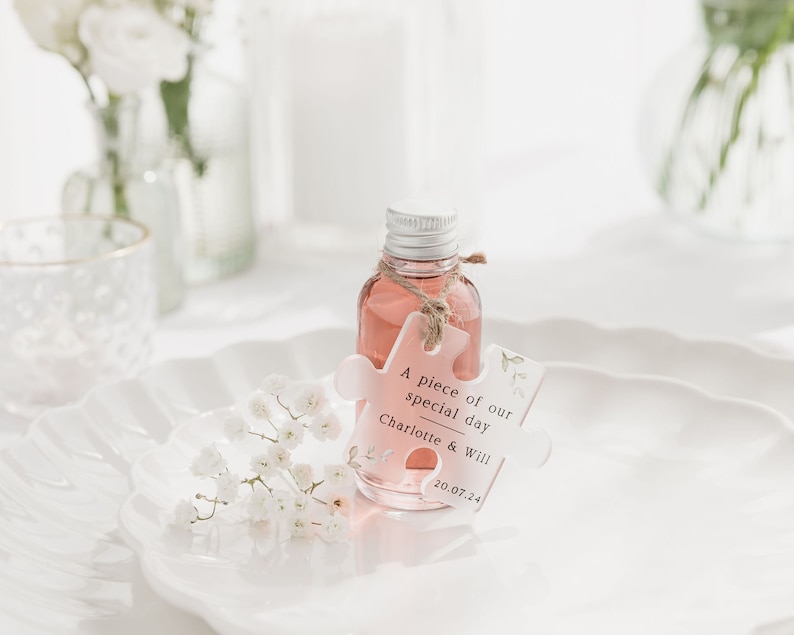 May include: A small clear bottle with a pink liquid and a white puzzle piece tag that says "A piece of our special day Charlotte & Will 20.07.24". The bottle is sitting on a white plate with white flowers.