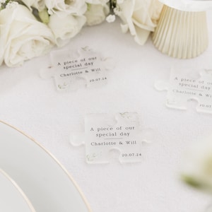 May include: Three clear plastic puzzle pieces with the text "A piece of our special day Charlotte & Will 20.07.24" printed on each piece. The puzzle pieces are on a white tablecloth with white flowers.