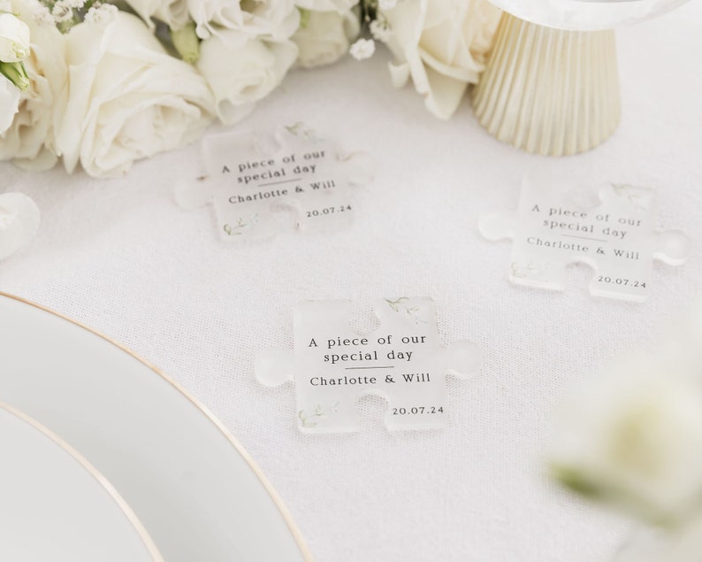 May include: Three clear plastic puzzle pieces with the text "A piece of our special day Charlotte & Will 20.07.24" printed on each piece. The puzzle pieces are on a white tablecloth with white flowers.