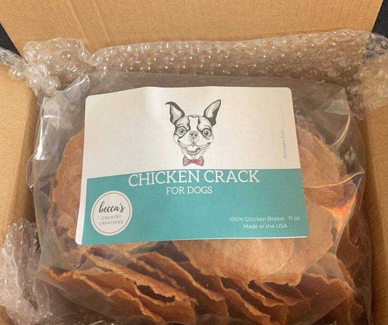 Puede incluir: Una bolsa de pl&aacute;stico transparente llena de golosinas de pollo deshidratado para perros, etiquetada como "Chicken Crack For Dogs". La etiqueta presenta un perro de dibujos animados con pajarita y el texto "100% Chicken Breast 11 oz Made in the USA."