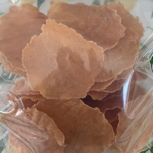 Puede incluir: Una bolsa de pl&aacute;stico transparente llena de una pila de finas patatas fritas de color marr&oacute;n claro, de forma irregular. Las patatas fritas tienen un aspecto ligeramente transl&uacute;cido y una superficie texturizada. La bolsa es parcialmente visible, mostrando que las patatas fritas est&aacute;n empaquetadas.