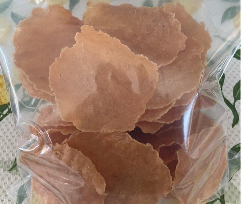 Puede incluir: Una bolsa de pl&aacute;stico transparente llena de una pila de finas patatas fritas de color marr&oacute;n claro, de forma irregular. Las patatas fritas tienen un aspecto ligeramente transl&uacute;cido y una superficie texturizada. La bolsa es parcialmente visible, mostrando que las patatas fritas est&aacute;n empaquetadas.