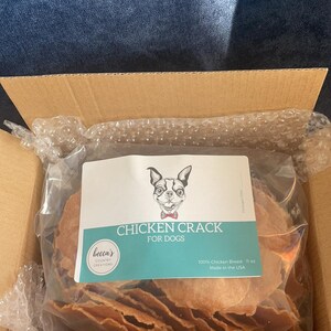 Puede incluir: Un paquete de golosinas para perros etiquetado como "Chicken Crack for Dogs" con una ilustraci&oacute;n de perro de dibujos animados. Las golosinas est&aacute;n hechas de 100% pechuga de pollo y pesan 312 gramos. El paquete est&aacute; dentro de una caja de cart&oacute;n.