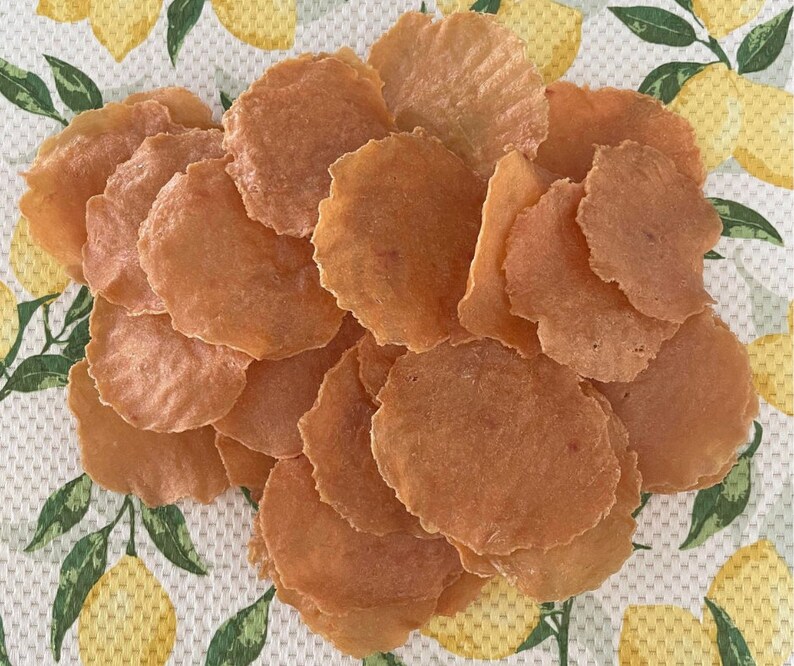 Puede incluir: Un mont&oacute;n de finas patatas fritas redondas de color marr&oacute;n claro. Las patatas fritas tienen un borde ligeramente ondulado y un aspecto transl&uacute;cido. Est&aacute;n dispuestas sobre un pa&ntilde;o blanco con un estampado de lim&oacute;n amarillo y hojas verdes.