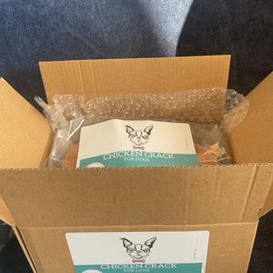 Puede incluir: Una caja de cart&oacute;n abierta que contiene un paquete de golosinas para perros. El paquete tiene una etiqueta con una ilustraci&oacute;n de perro y el texto "CHICKEN CRACK FOR DOGS". La caja est&aacute; sobre una superficie azul.