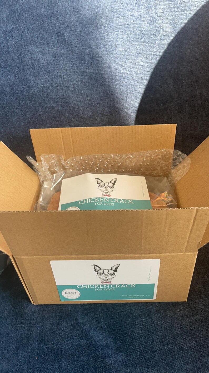 Puede incluir: Una caja de cart&oacute;n abierta que contiene un paquete de golosinas para perros. El paquete tiene una etiqueta con una ilustraci&oacute;n de perro y el texto "CHICKEN CRACK FOR DOGS". La caja est&aacute; sobre una superficie azul.