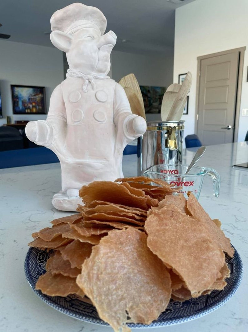 Puede incluir: Una estatua de cerdo chef de cer&aacute;mica blanca est&aacute; junto a un plato de comida crujiente de color marr&oacute;n dorado. Tambi&eacute;n se ven una taza medidora Pyrex y un soporte de utensilios de metal con cucharas de madera. El plato tiene un borde con dibujos azules y blancos.