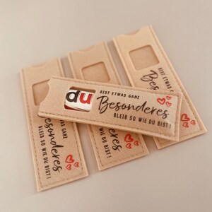 Könnte beinhalten: Mehrere beige Filzhüllen mit rechteckigem Ausschnitt, die jeweils eine kleine Schokoriegel enthalten. Die Hüllen tragen das Wort "du" und das Wort "Besonderes", sowie rote Herzgrafiken. Der Text "BIST ETWAS GANZ" und "BLEIB SO WIE DU BIST!" ist ebenfalls aufgedruckt.