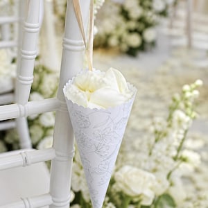 May include: A white chair adorned with a cone-shaped paper holder filled with white flower petals. The holder, patterned with flowers, is suspended by a gold ribbon. The background is filled with white floral arrangements.