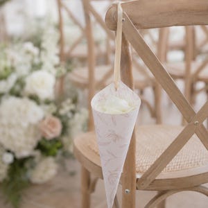 May include: A paper cone filled with white flower petals, featuring a delicate light pink floral line drawing pattern. The cone is suspended by a thin, light-coloured ribbon attached to the back of a light brown wooden cross-back chair.