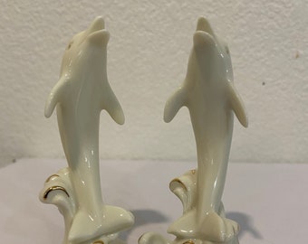 May include: A pair of white porcelain dolphin figurines with gold accents. The dolphins are leaping out of the water, which is depicted in a stylized wave pattern.