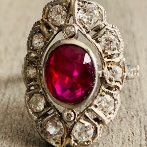 May include: A vintage ring with a large oval ruby surrounded by a halo of smaller diamonds. The ring is set in silver and has a delicate, intricate design.