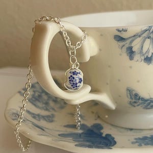 May include: A silver chain necklace with a blue and white porcelain pendant featuring a floral design. The pendant is round and hangs from the chain.