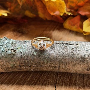 May include: A gold wire wrapped ring with a white ceramic bead featuring a floral pattern. The ring is resting on a piece of wood with a brown bark texture.