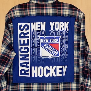 May include: A blue and white plaid flannel shirt with a blue patch on the back. The patch features the New York Rangers logo and the words "New York Rangers Hockey".
