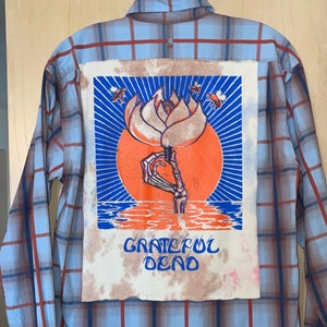 Puede incluir: Una camisa de franela a cuadros azules y rojos con un parche tie-dye de Grateful Dead en la espalda. El parche presenta una mano esquelética sosteniendo una flor con un sol y agua detrás.