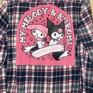 Puede incluir: Camisa de franela de manga larga con botones y un estampado de cuadros en azul marino, verde y rosa. La espalda presenta un parche rosa con el texto "My Melody & Kuromi" y las palabras "Pretty & Bold" con personajes de dibujos animados.