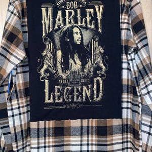 May include: A long-sleeved flannel shirt with a black patch featuring Bob Marley's image and the words "Marley Rebel Music Legend" in gold lettering. The shirt has a brown, black, and white plaid pattern.